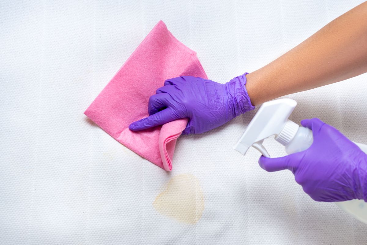 Mano con guantes morados tratando una mancha en una cama con un trapo rosa