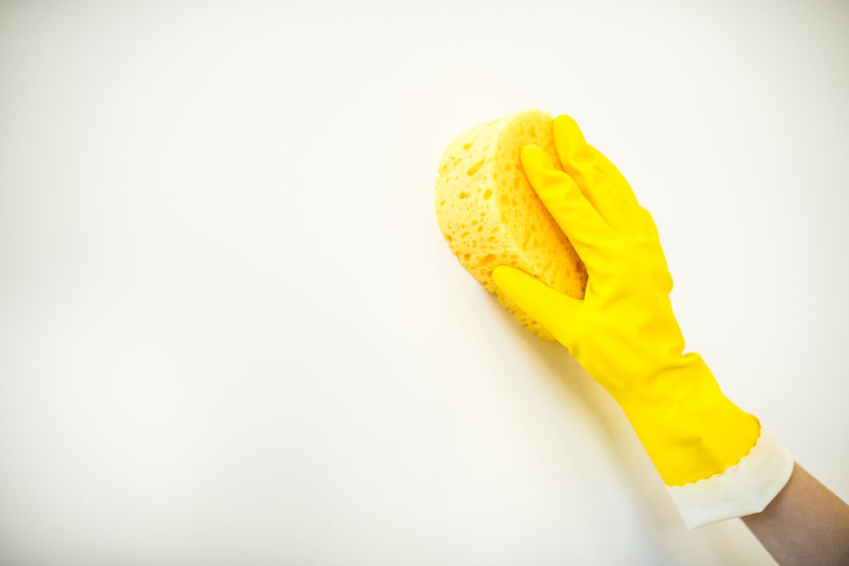 Mano con guante de goma amarillo limpiando una pared blanca con una esponja amarilla
