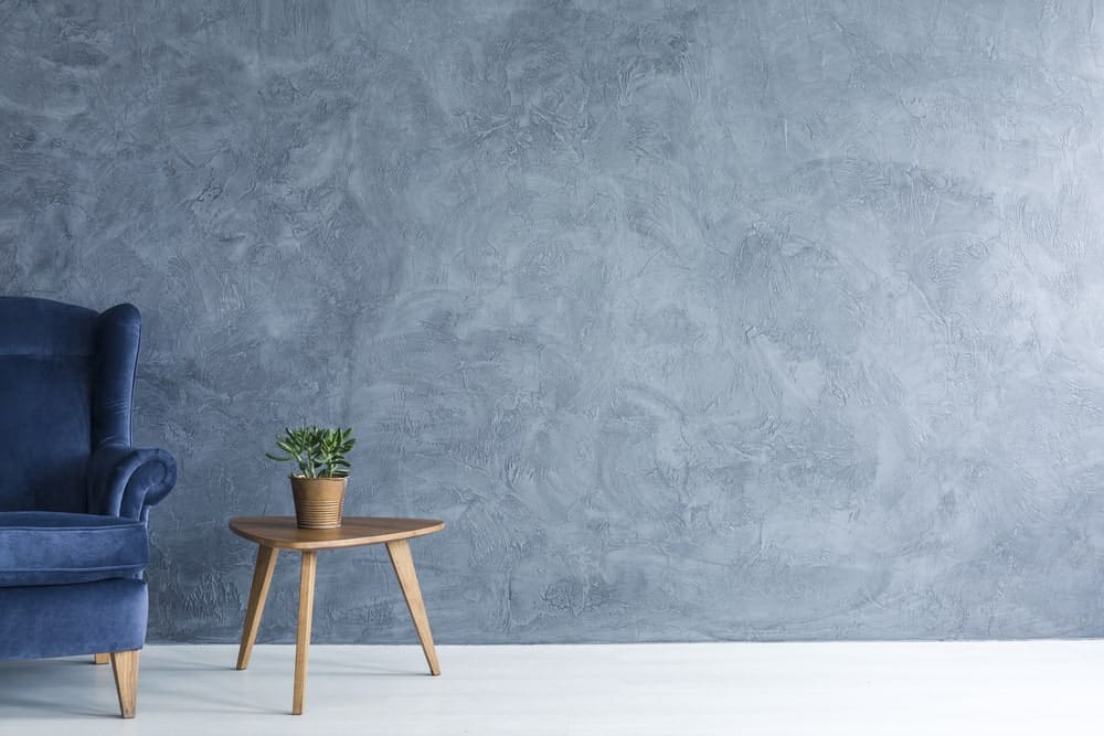 Paredes minimalistas azuladas con un sillón de terciopelo azul y una mesa auxiliar de madera con una planta en maceta.