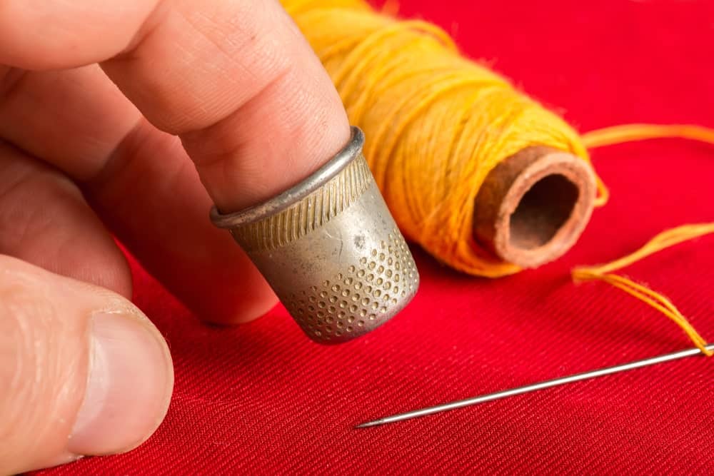 Primer plano del dedo de punta con dedal junto a un hilo y una aguja amarillos sobre fondo de tela roja.