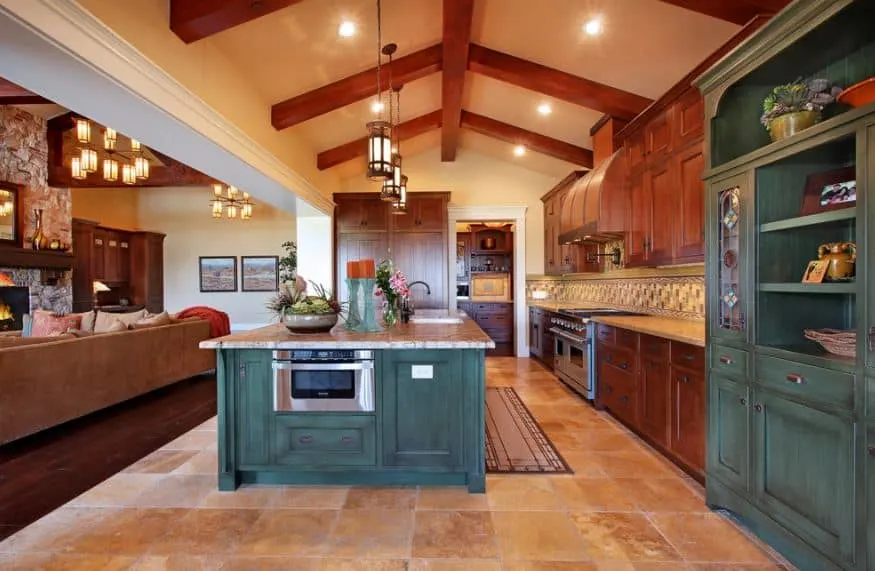 La cocina de estilo artesano tiene un techo catedral con vigas de madera vistas que se funden con los armarios. Una isla central tiene un horno empotrado con cajones y armarios de madera, y un acabado teñido de verde.