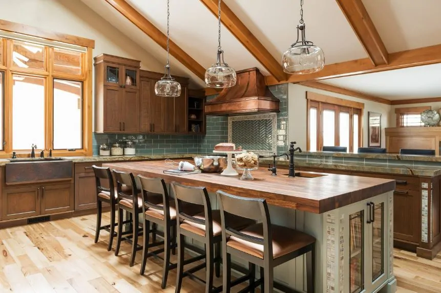 Las lámparas colgantes cuelgan del techo blanco de esta cocina con vigas de madera a la vista que reflejan los marcos de las altas ventanas. Estas ventanas proporcionan iluminación natural a los tonos de madera oscura de la isla de cocina y los armarios de la península. 