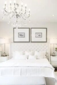 La tradicional cama con mechón se centra en la pared de este tranquilo dormitorio blanco. Mesillas de noche a juego con lámparas de cristal flanquean ambos lados de la cama y encima cuelga un cuadro de arte de la naturaleza.