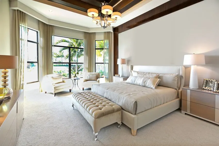 Este dormitorio principal cuenta con un elegante juego de cama sobre el suelo de moqueta blanca. La habitación ofrece una zona de estar cerca de las ventanas. También tiene un impresionante techo iluminado por una elegante lámpara de techo.