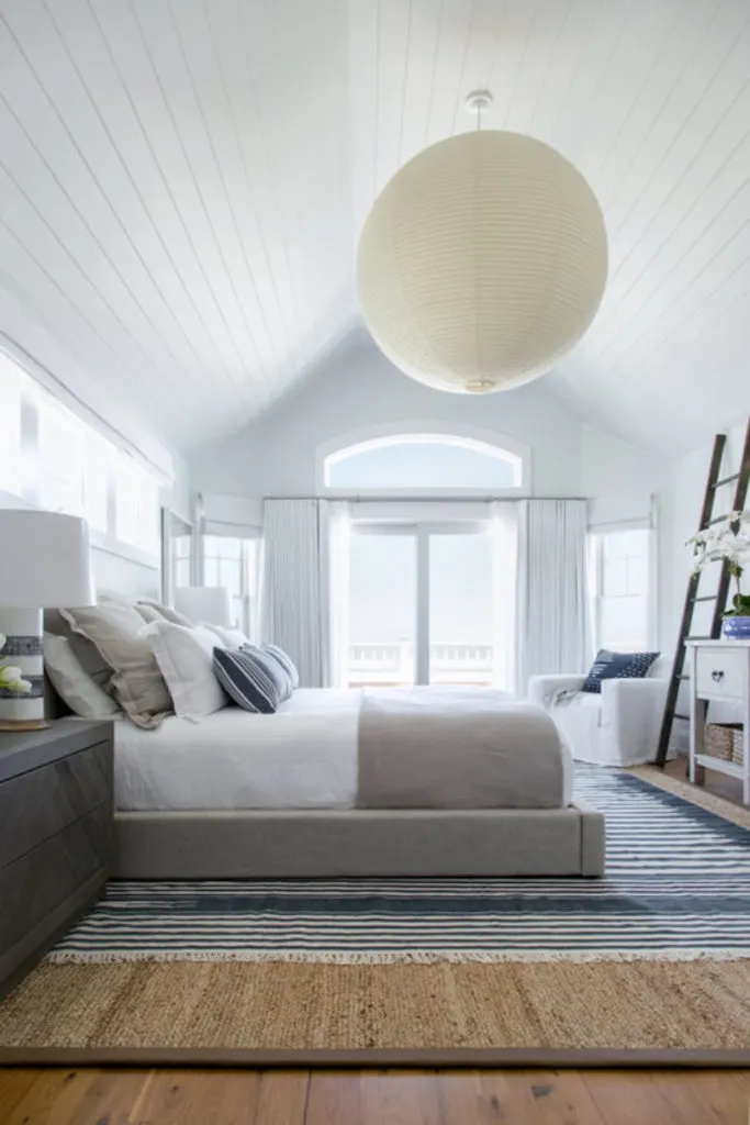 Dormitorio principal con suelo de madera noble rematado por una gran alfombra rodeada de paredes blancas y techo con una enorme iluminación colgante.