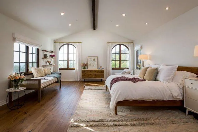 Amplio dormitorio principal con suelo de madera noble rematado por una gran alfombra, paredes blancas y techo abovedado con una viga vista.