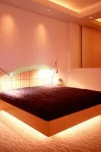 Un vistazo más de cerca a la impresionante cama a medida de este dormitorio principal, iluminada con luz brillante, junto con mesillas de noche empotradas. La habitación tiene techo de bandeja y suelo de moqueta.